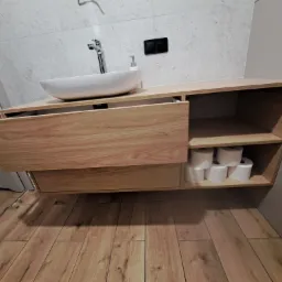 Nowoczesna łazienka z drewnianą szafką pod umywalką nablatową i zapasem papieru toaletowego na p&oacute;łce.