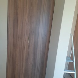 KJK Meble Krzysztof Krzysztofowicz - Dwu-drzwiowa szafa z okleiną imitującą drewno orzechowe, z minimalistycznym, podłużnym uchwytem, ustawiona przy ścianie w odcieniach beżu, z widoczną drabinką malarską.
