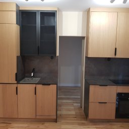 KJK Meble Krzysztof Krzysztofowicz - Jasna kuchnia z drewnianymi frontami, czarnymi akcentami i szarym blatem. Widoczny zlew, płyta grzewcza i piekarnik. Podłoga drewniana. Ujęcie proste, bez dekoracji.