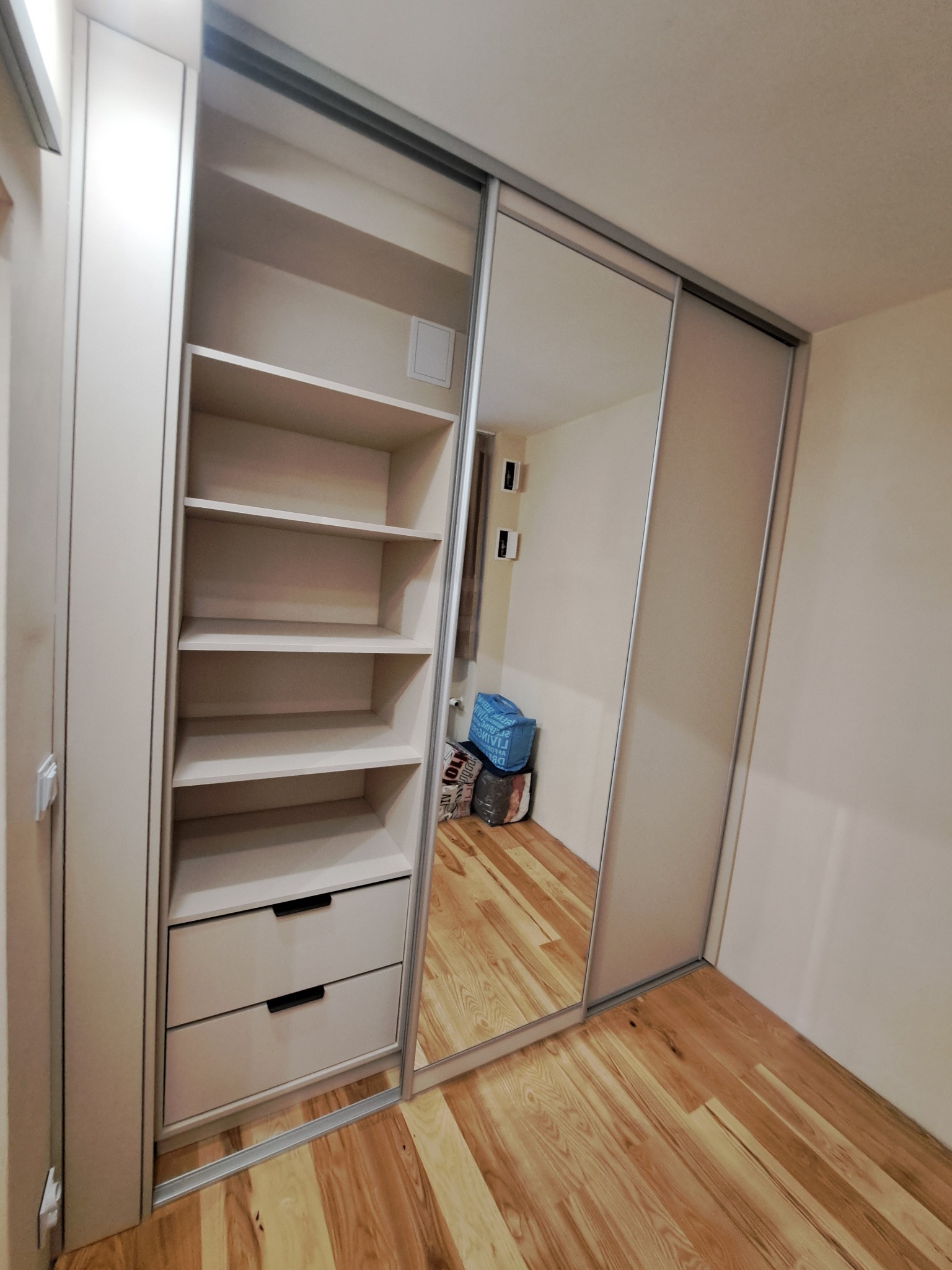 Jasna szafa wnękowa z przesuwnymi drzwiami z lustrem, widoczne półki i dwie szuflady, na podłodze parkiet.