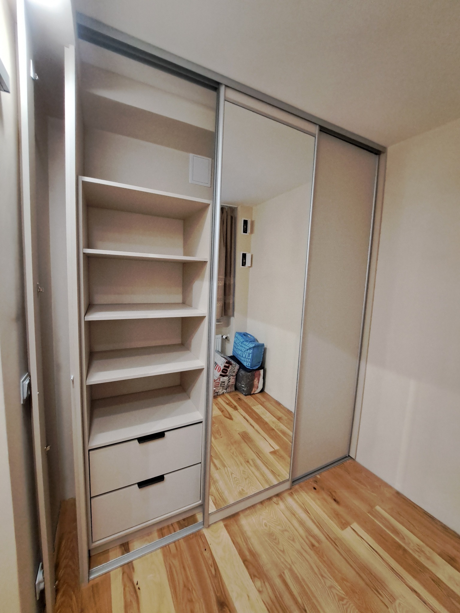 Beżowa szafa przesuwna z lustrem i otwartą częścią z półkami i dwiema szufladami, na drewnianej podłodze.