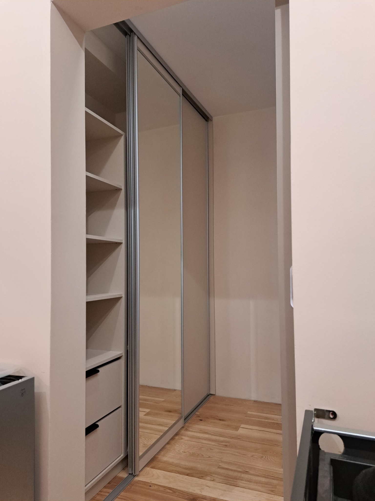 Wnętrze wnęki z otwartą szafą z przesuwnymi, lustrzanymi drzwiami, widoczne półki i dwie szuflady w jasnej kolorystyce, drewniana podłoga.