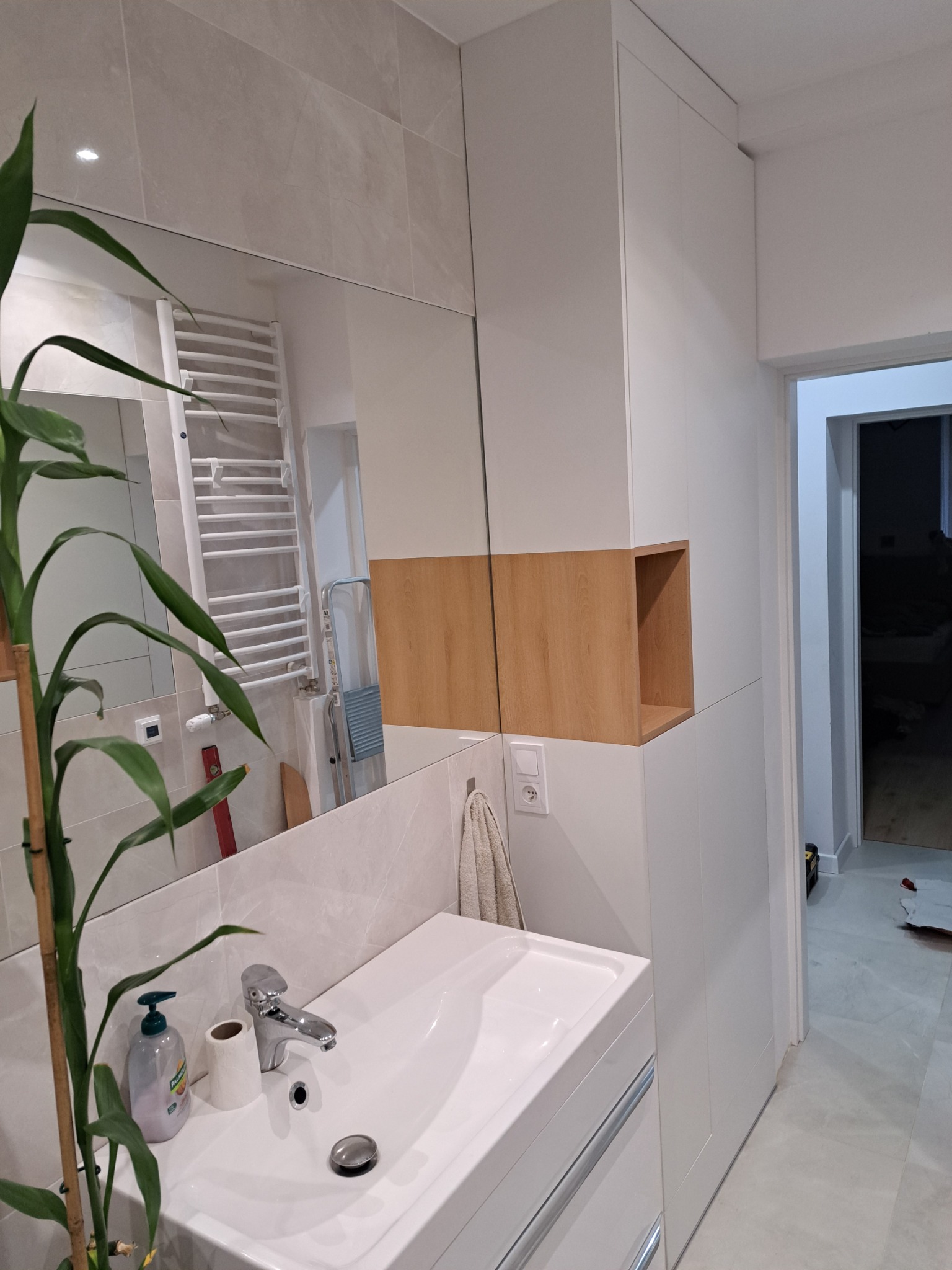 Nowoczesna łazienka z białą szafą wnękową z drewnianą wnęką, umywalką nablatową i bambusową rośliną w rogu.