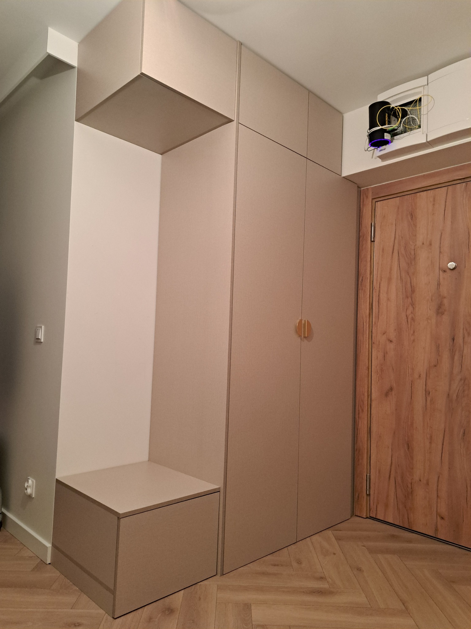 Beżowa zabudowa w przedpokoju z siedziskiem, szafą i otwartą przestrzenią na wieszaki, obok drzwi wejściowe z drewnopodobnym wzorem, podłoga ułożona w jodełkę.