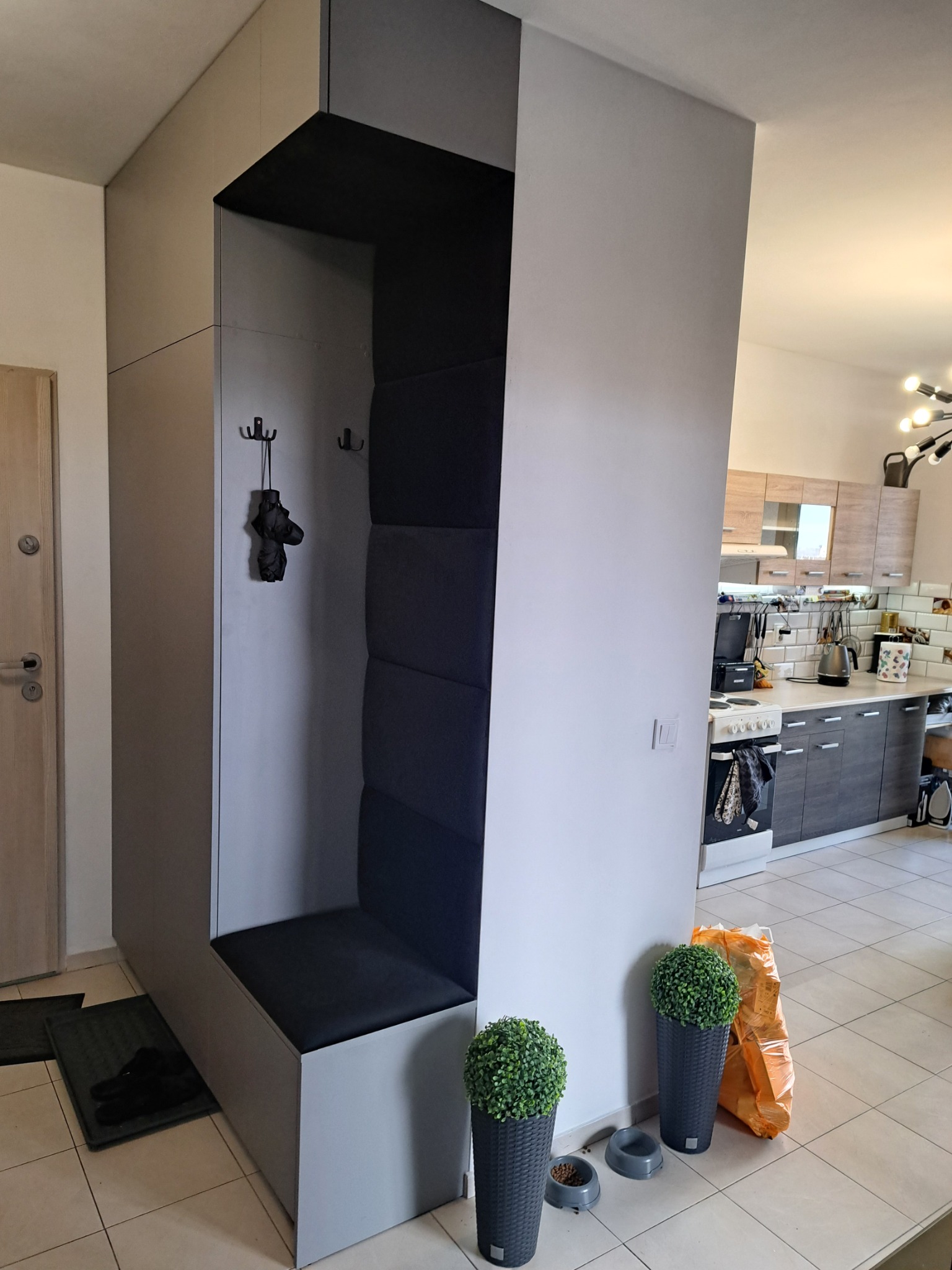 Szara zabudowa w przedpokoju z siedziskiem i wieszakami, tapicerowany panel na ścianie, widok na kuchnię w tle.