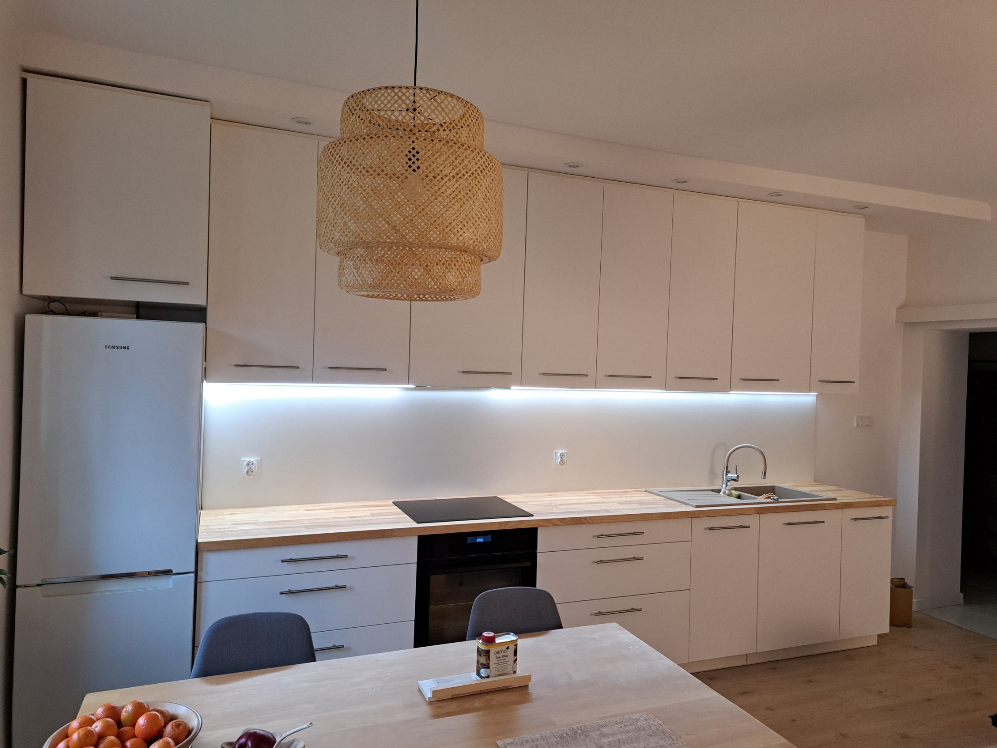 Nowoczesna kuchnia z białymi szafkami, drewnianym blatem, plecioną lampą i widocznym na pierwszym planie stołem z owocami.