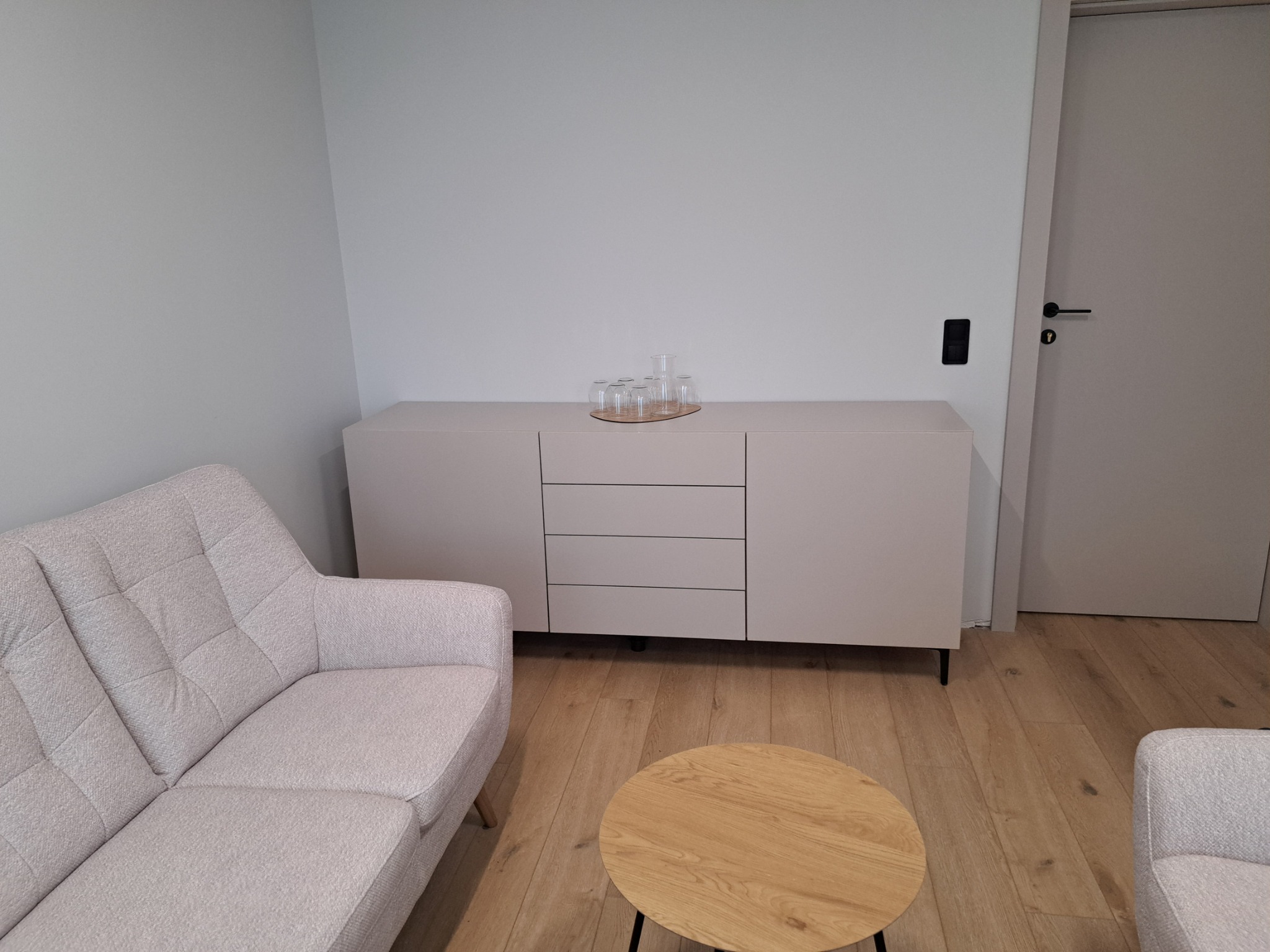 Jasnoszara komoda z trzema szufladami i szafkami, na której stoi zestaw szklanek, w minimalistycznym salonie z drewnianą podłogą i tapicerowaną sofą w odcieniach szarości.