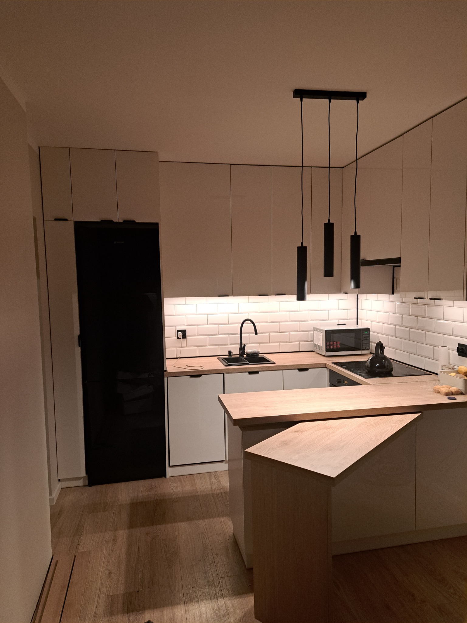 Nowoczesna kuchnia z białymi frontami i drewnianym blatem w Police, oświetlona wiszącymi lampami i podświetleniem LED pod szafkami, z czarną lodówką w zabudowie.