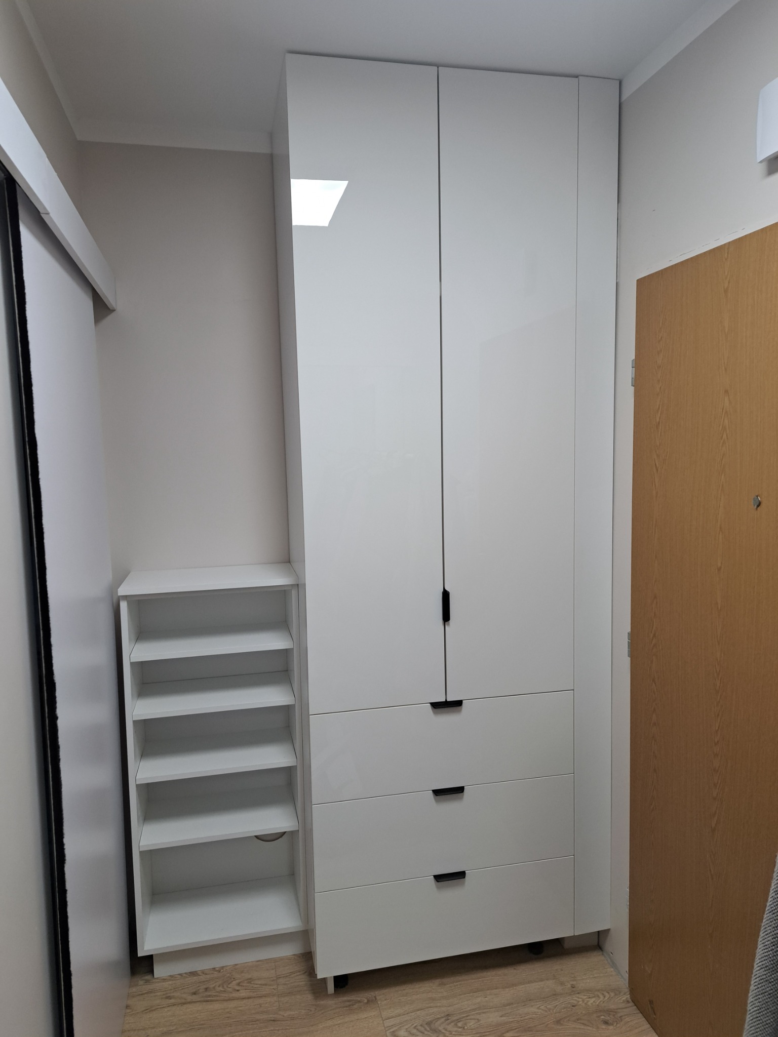 Biała, wysoka szafa z połyskiem, trzema szufladami i czarnymi uchwytami, stojąca obok białego regału z półkami, w jasnym wnętrzu.