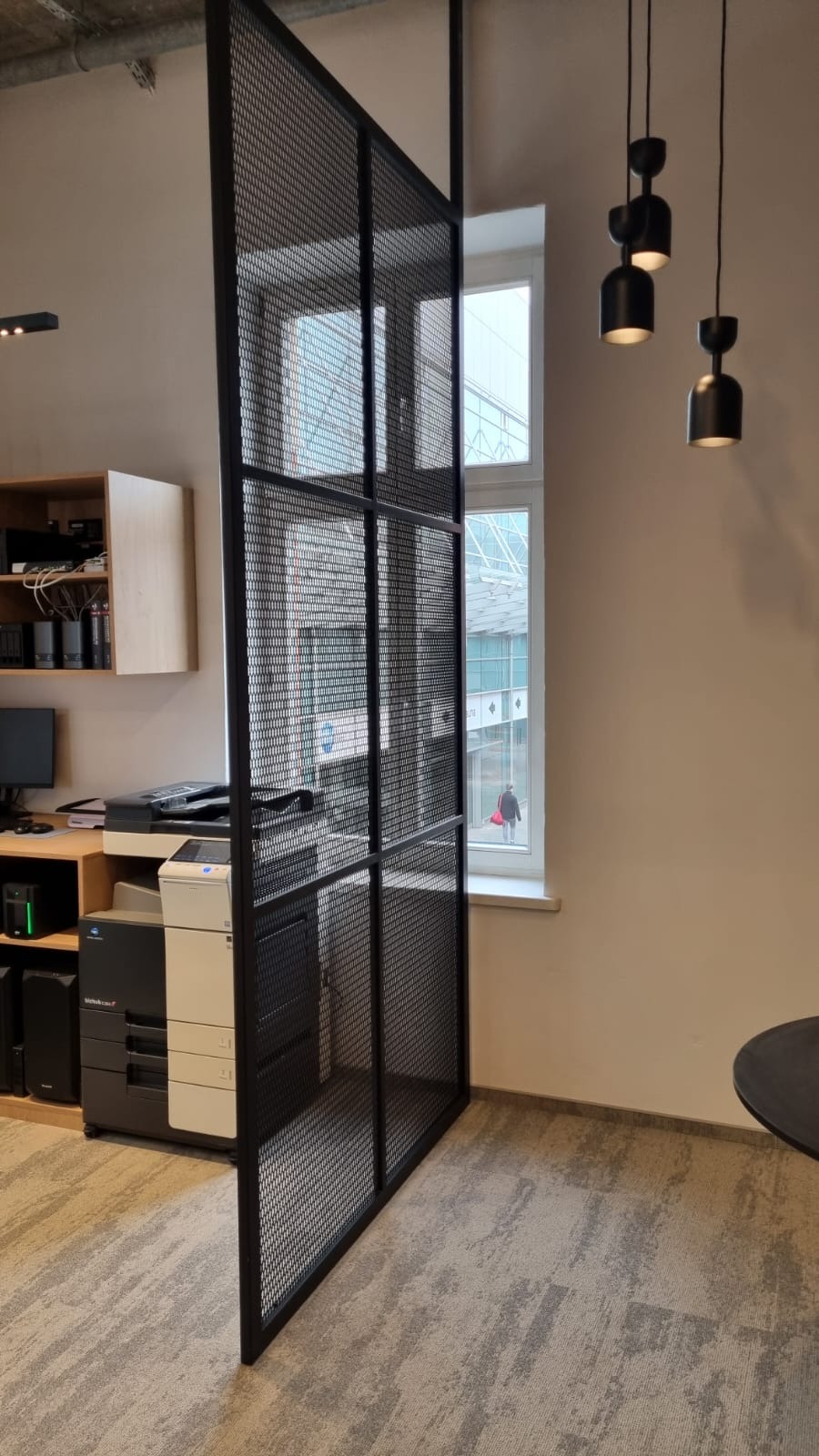 Ażurowa, metalowa ścianka działowa w biurze, z widocznym sprzętem biurowym w tle i trzema lampami wiszącymi.