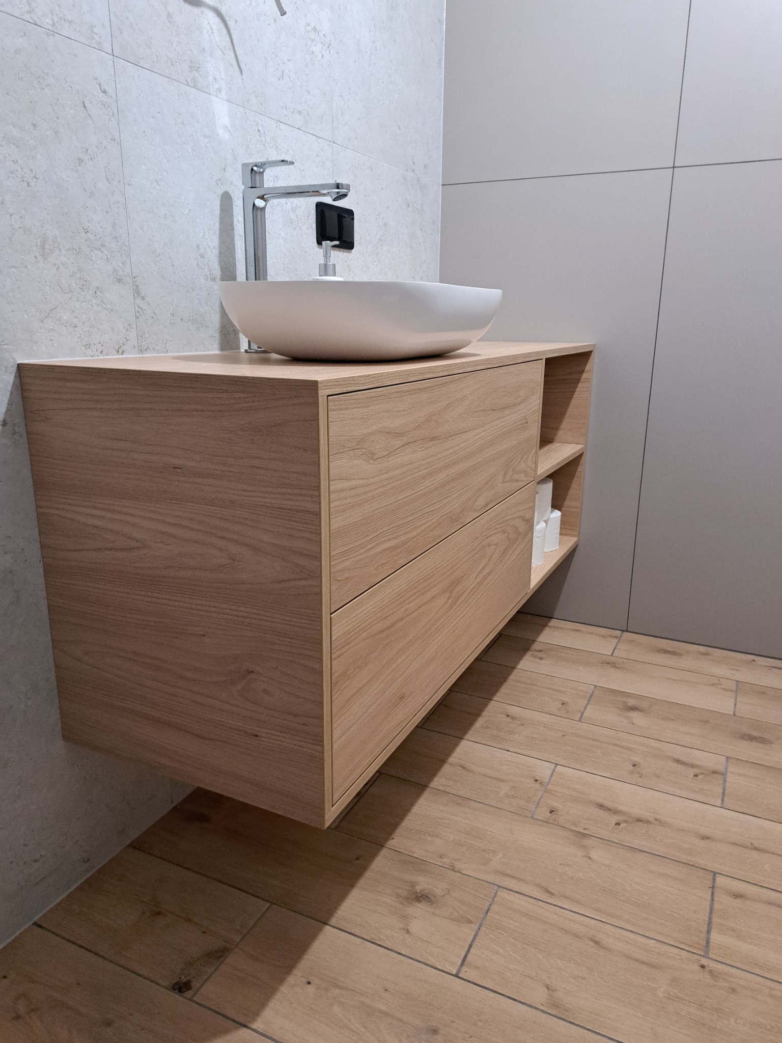 Drewniana szafka pod umywalkę z dwoma szufladami i otwartą półką boczną, na której widoczne są zwinięte ręczniki papierowe; umywalka nablatowa w kształcie misy i nowoczesna bateria.