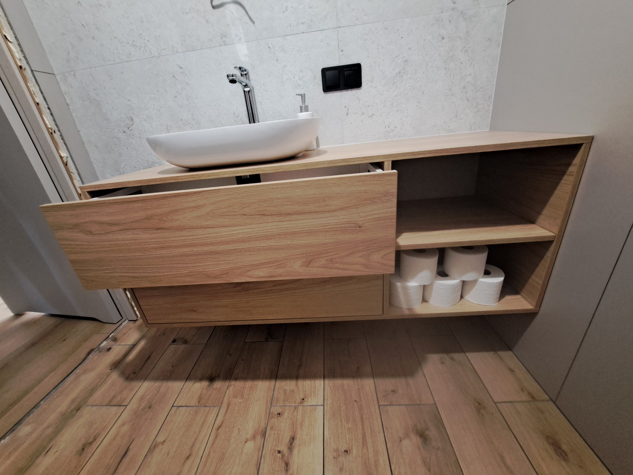 Nowoczesna łazienka z drewnianą szafką pod umywalką nablatową i zapasem papieru toaletowego na półce.