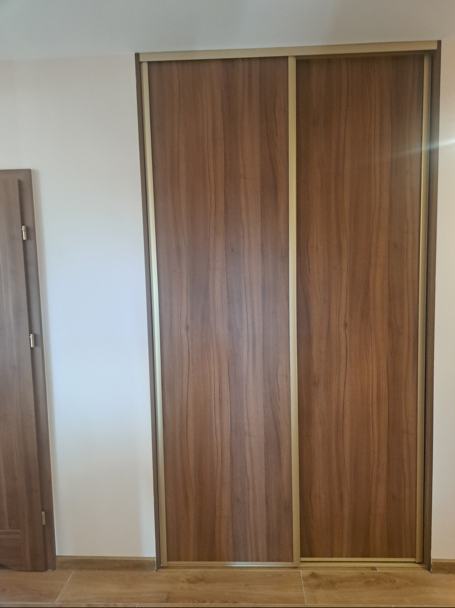 Dwu-drzwiowa szafa wnękowa z przesuwnymi drzwiami o wykończeniu imitującym drewno orzechowe, z delikatnymi złotymi ramkami, widoczna na tle białej ściany i fragmentu podłogi.