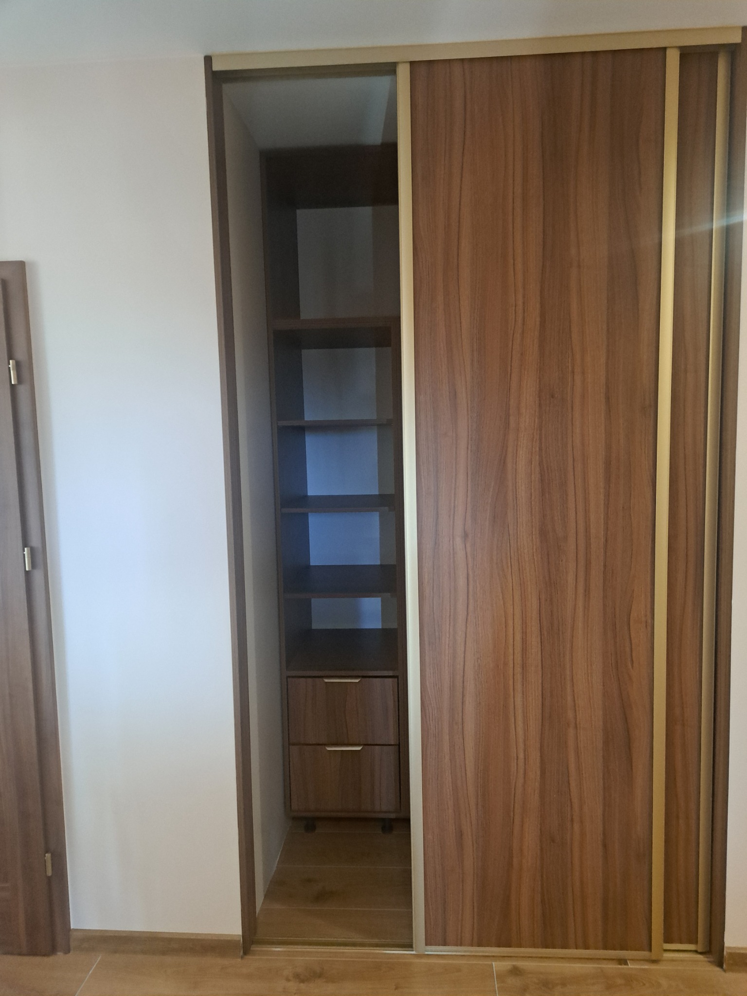 Wnętrze szafy wnękowej z przesuwnymi drzwiami o wykończeniu drewnopodobnym, z widocznymi półkami i dwiema szufladami, oświetlonej światłem dziennym.