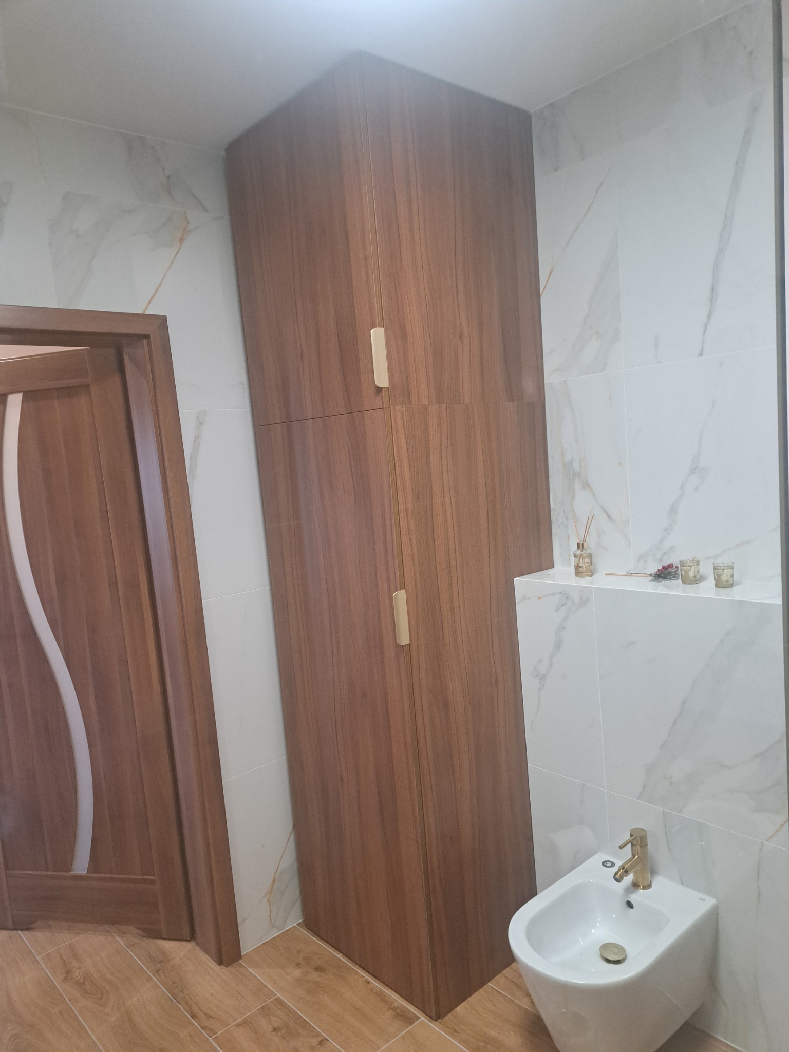Wysoka, drewniana szafa w łazience z marmurowymi płytkami, obok drzwi i bidetu z złotą armaturą.