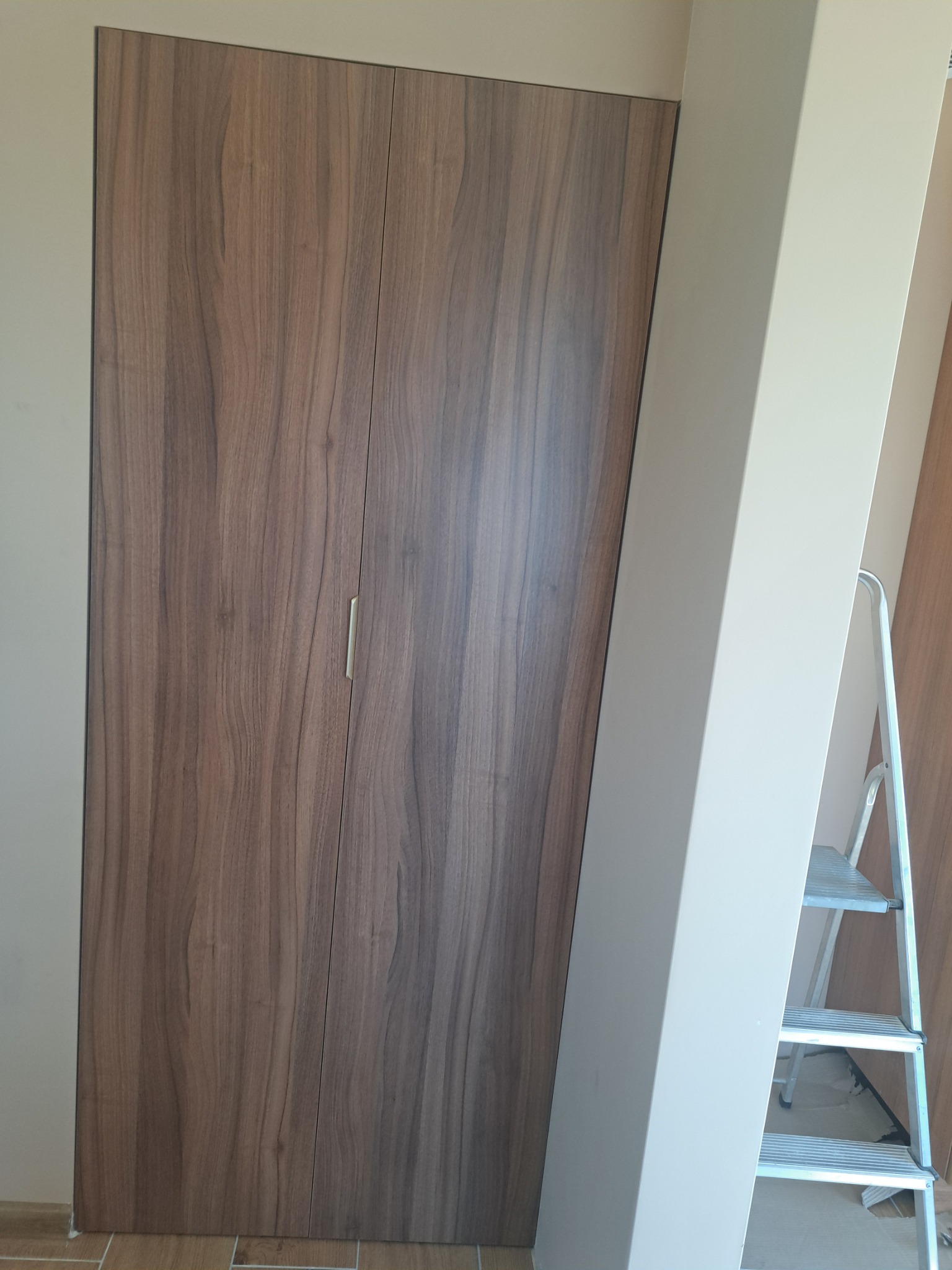 Dwu-drzwiowa szafa z okleiną imitującą drewno orzechowe, z minimalistycznym, podłużnym uchwytem, ustawiona przy ścianie w odcieniach beżu, z widoczną drabinką malarską.