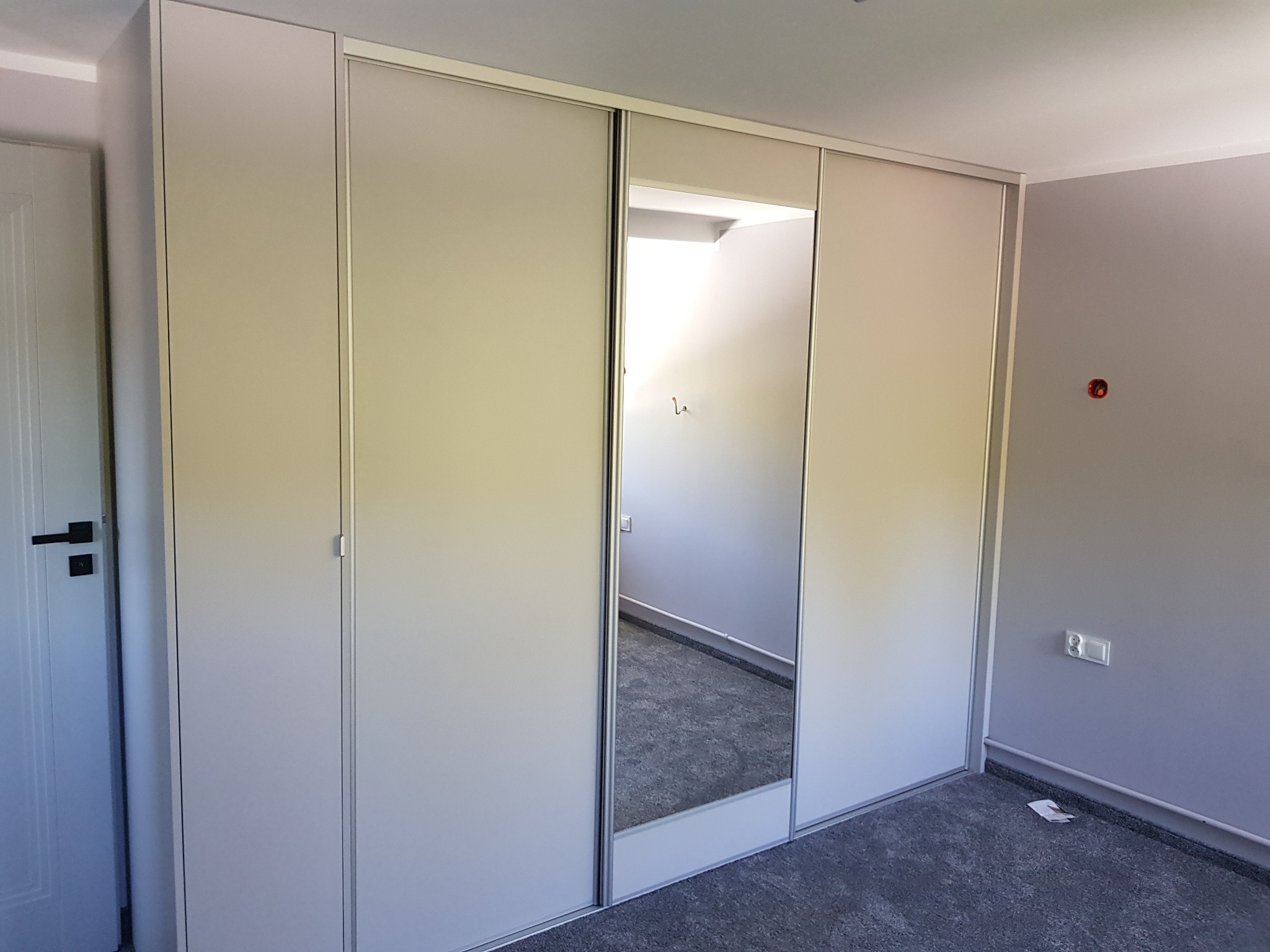 Jasna szafa wnękowa z przesuwnymi drzwiami i lustrem, odbijającym wnętrze garderoby. Minimalistyczny design, szare ściany, widoczna instalacja elektryczna.