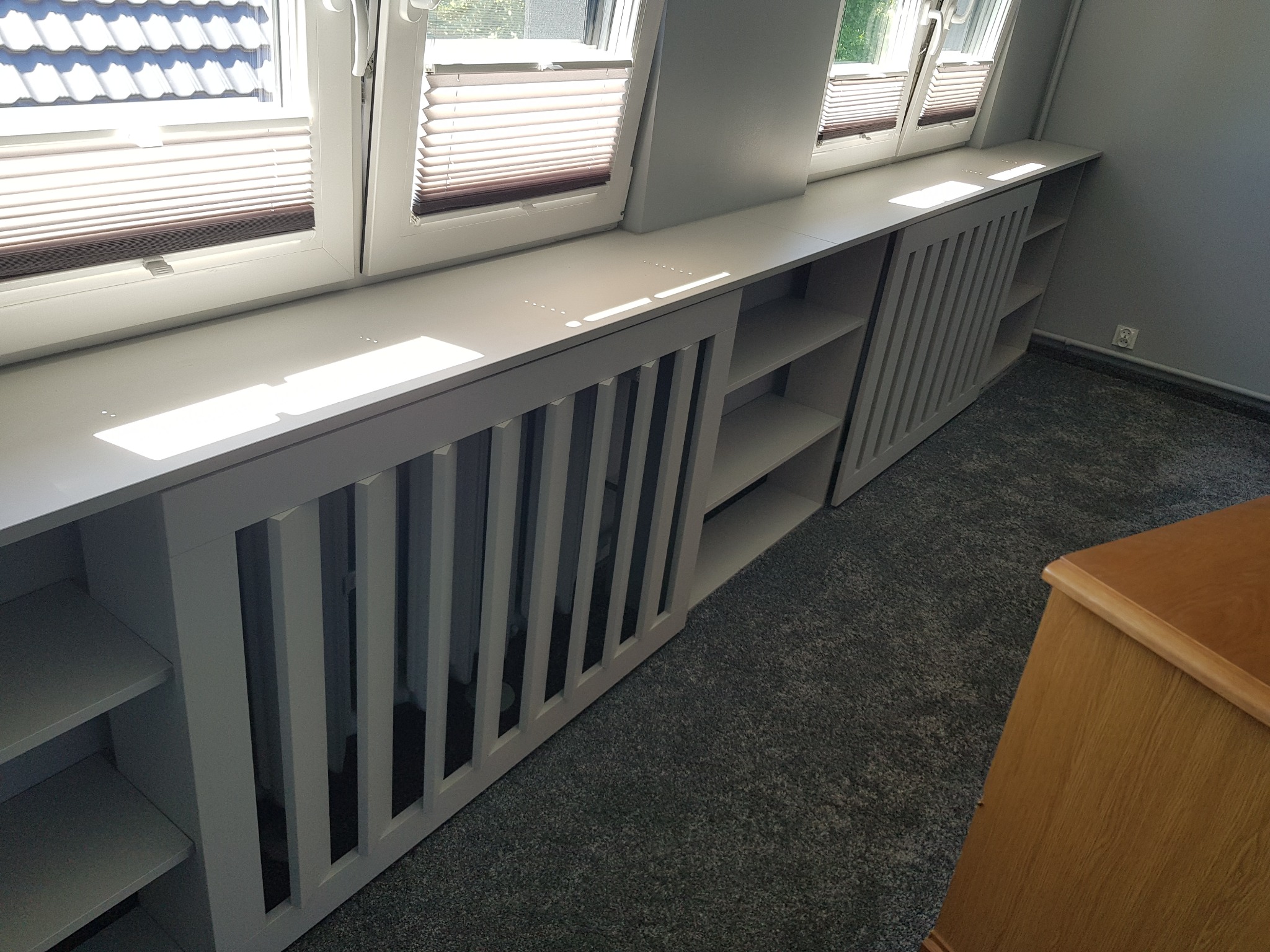 Biała zabudowa okienna z półkami i ażurowymi osłonami grzejników, dopasowana do skosów okiennych. Nowoczesny design i funkcjonalność w jednym.