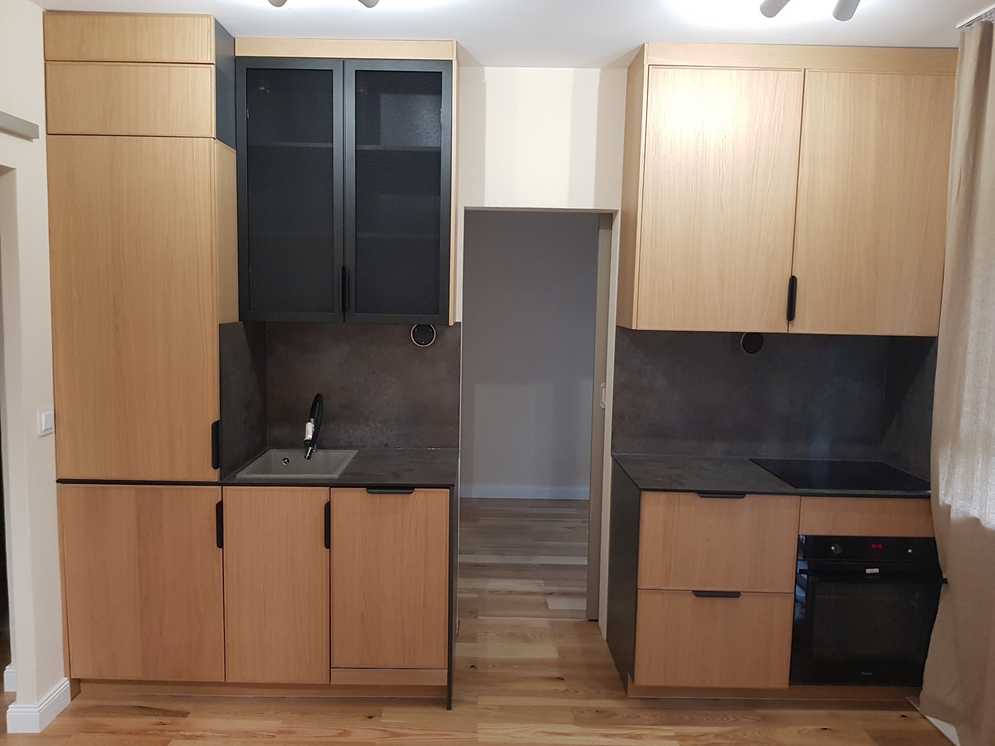 Jasna kuchnia z drewnianymi frontami, czarnymi akcentami i szarym blatem. Widoczny zlew, płyta grzewcza i piekarnik. Podłoga drewniana. Ujęcie proste, bez dekoracji.