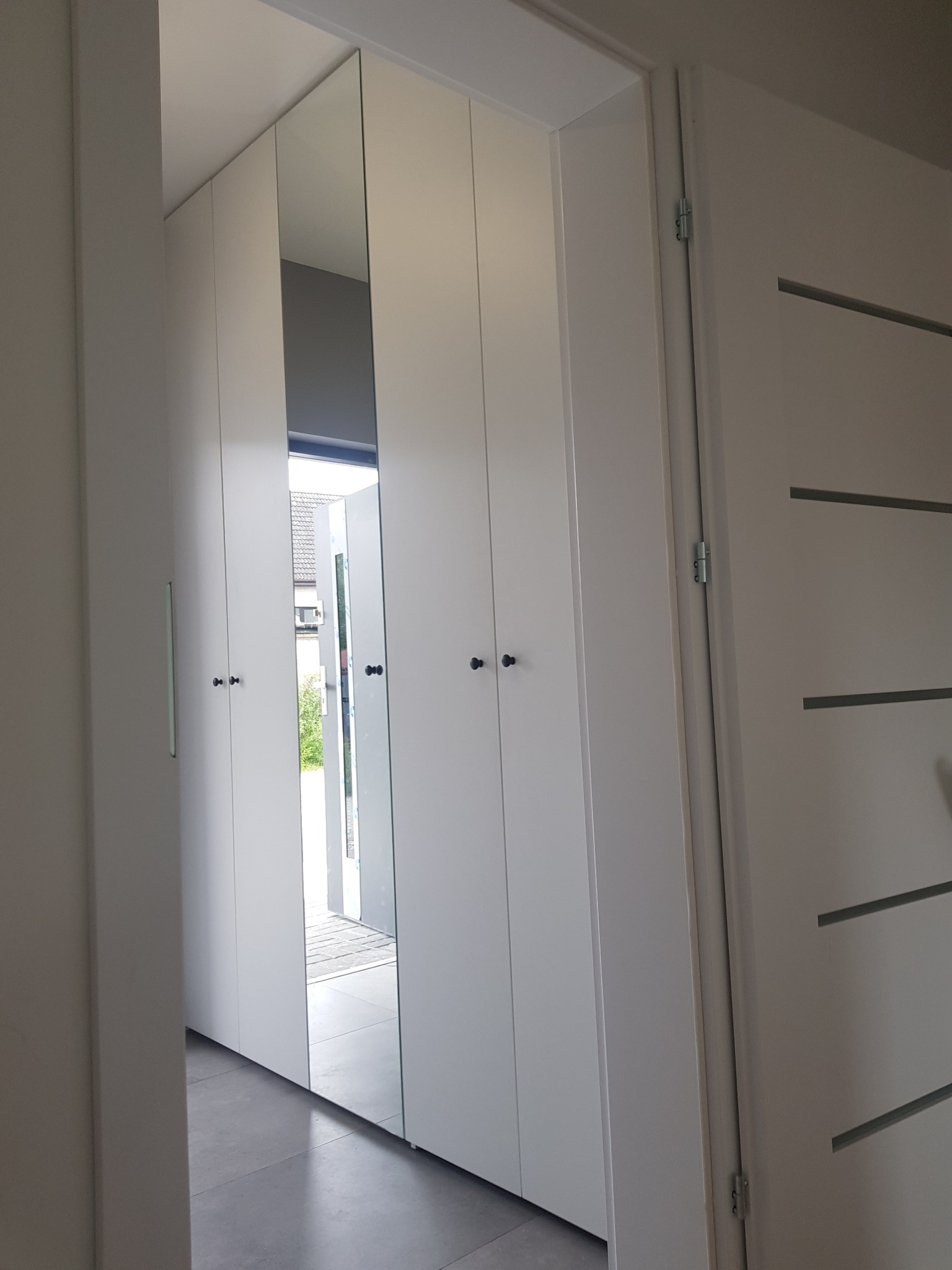 Biała szafa wnękowa z lustrem w przedpokoju, widok z boku, czarne okrągłe uchwyty, szara podłoga, drzwi wejściowe z poziomymi wstawkami w tle.