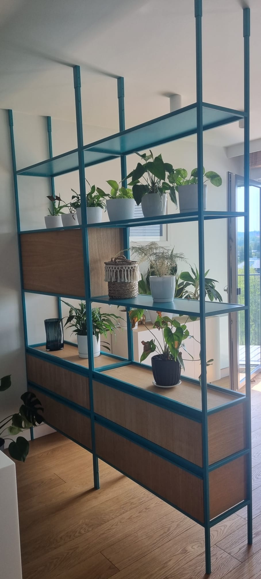 Turkusowy regał z drewnianymi panelami, wypełniony roślinami doniczkowymi, tworzący ażurową ściankę działową w jasnym wnętrzu z parkietem.