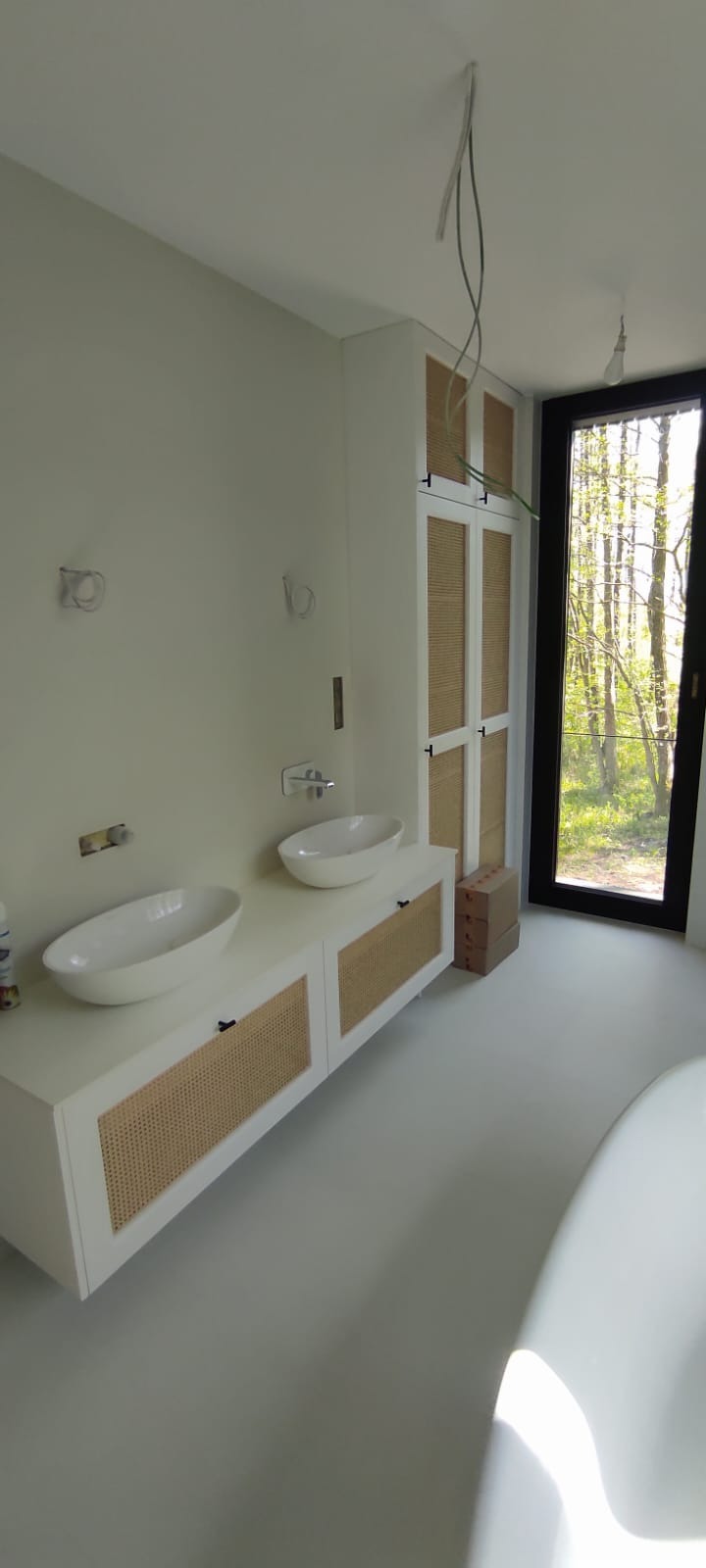 Biała szafka łazienkowa z rattanowymi frontami pod umywalki nablatowe, obok szafa w tym samym stylu. Widok na okno z zielenią. Wnętrze w neutralnych barwach.