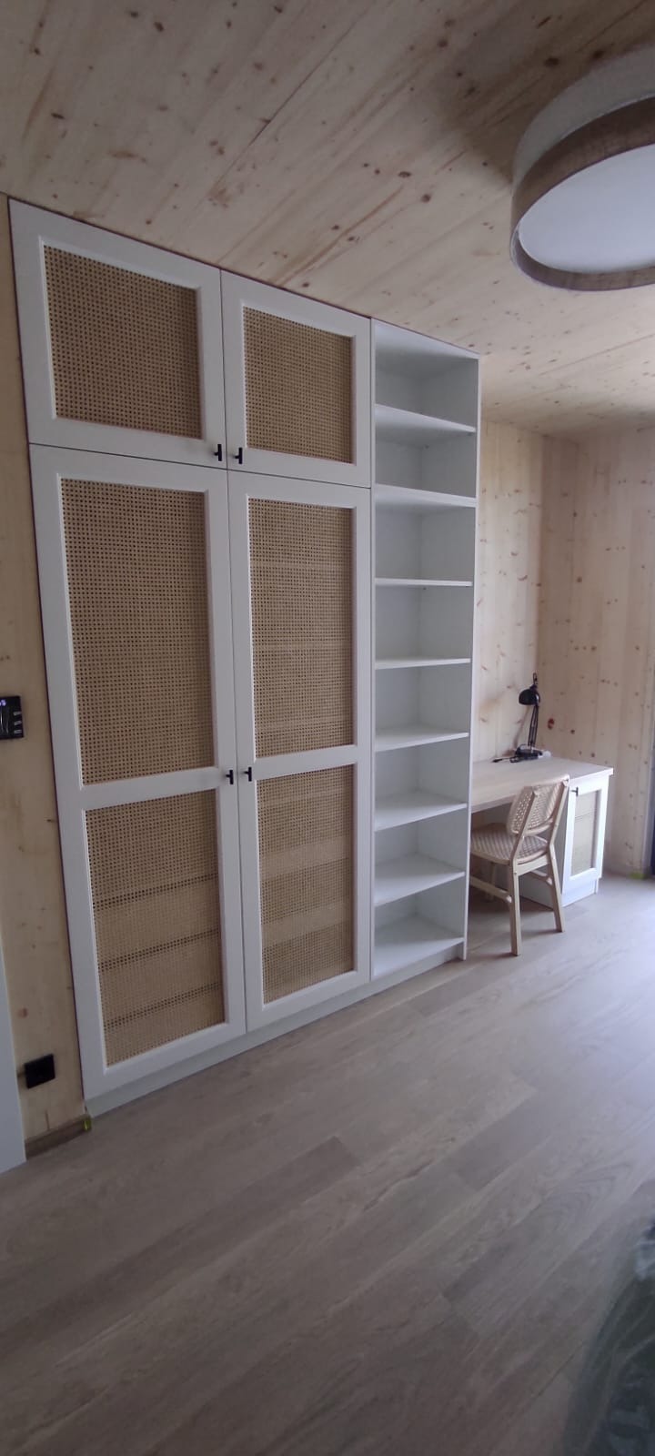 Biała szafa wnękowa z rattanowymi frontami, połączona z regałem i biurkiem w pokoju wykończonym drewnem. Minimalistyczny design, naturalne materiały.