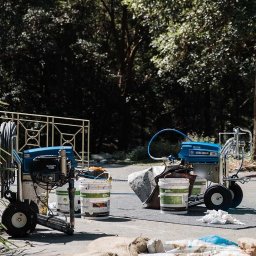 Profi - Dwa agregaty malarskie Ultra Max II 795 z wiaderkami farby Roof Protect na szarym podłożu, w tle zieleń drzew. Sprzęt gotowy do natryskowego malowania.