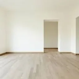 Jasne, puste wnętrze po remoncie z parkietem, widok na ogród przez duże okno i dwa otwory drzwiowe. Świeżo pomalowane ściany i listwy przypodłogowe.