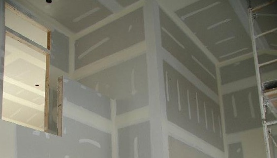 Wnętrze w trakcie prac wykończeniowych: ściany i sufit pokryte płytami gipsowo-kartonowymi, widoczny fragment ramy okiennej i rusztowania. Przygotowanie do malowania.