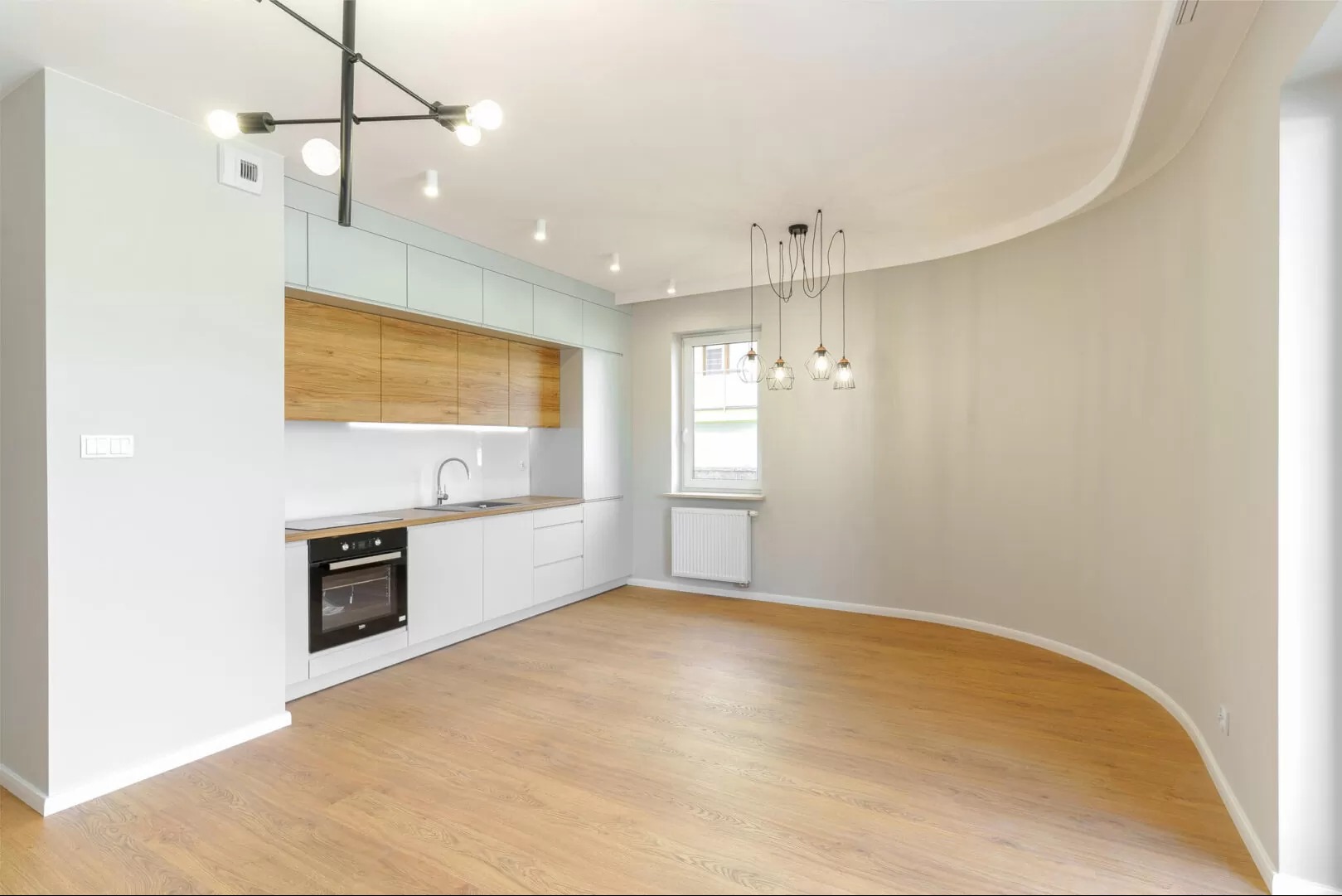 Jasne wnętrze po remoncie: białe ściany, drewniana podłoga, nowoczesna kuchnia z drewnianymi akcentami i designerskie lampy. Widok na odnowione mieszkanie.