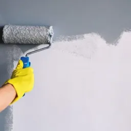 Wałek malarski w trakcie pracy, nakładający szarą farbę na białą ścianę, widoczna dłoń w żółtej rękawicy ochronnej.