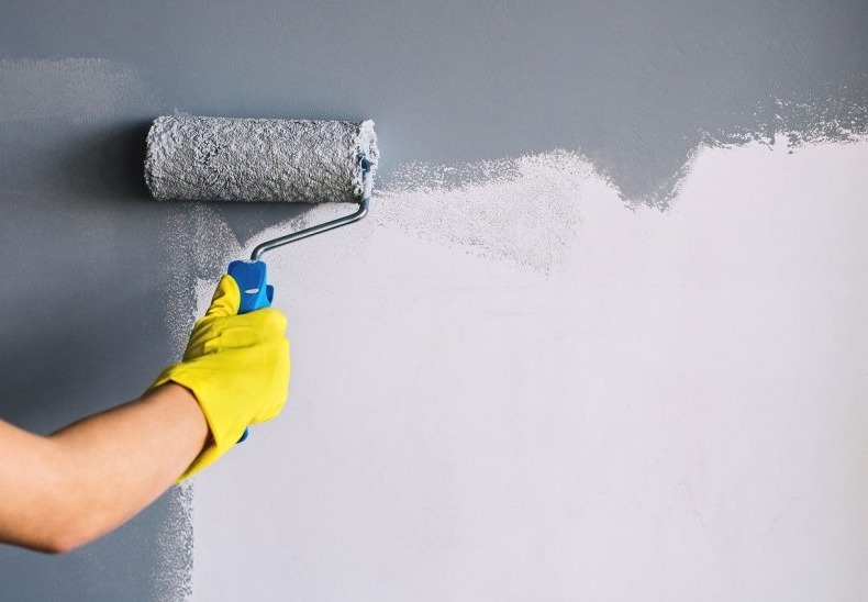 Wałek malarski w trakcie pracy, nakładający szarą farbę na białą ścianę, widoczna dłoń w żółtej rękawicy ochronnej.