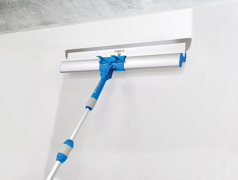 Narzędzie do rozprowadzania masy szpachlowej z teleskopową rączką marki Storch, przymocowane do białej ściany, widoczny świeżo nałożony fragment masy.