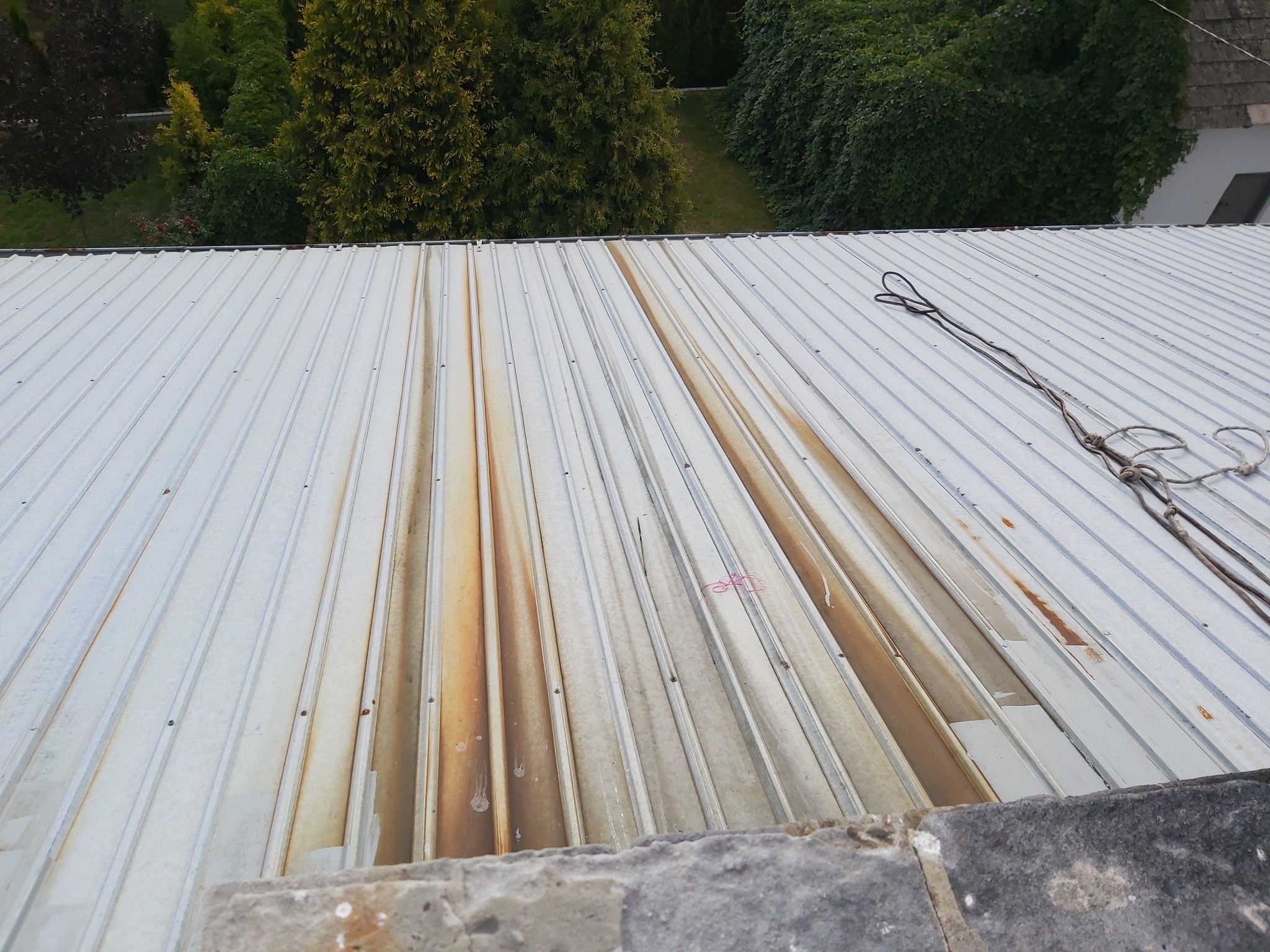 Metalowy dach z widocznymi smugami zabrudzeń i śladami po czyszczeniu, lina zabezpieczająca na dachu, w tle zieleń drzew