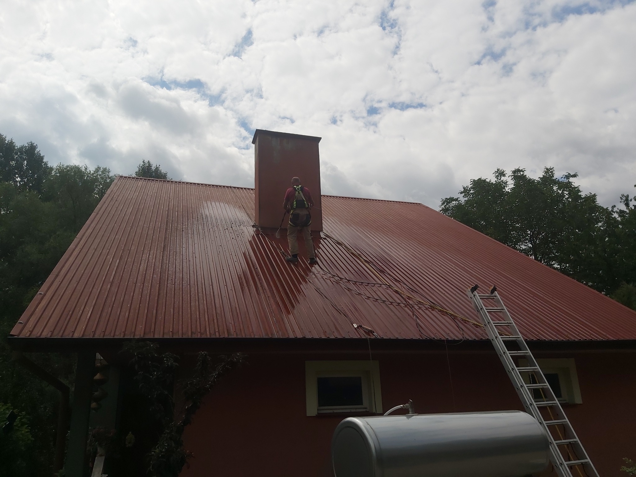 Pracownik w uprzęży na mokrym, czerwonym dachu z blachy trapezowej, czyści go za pomocą lancy ciśnieniowej. W tle komin i drabina oparta o dach.