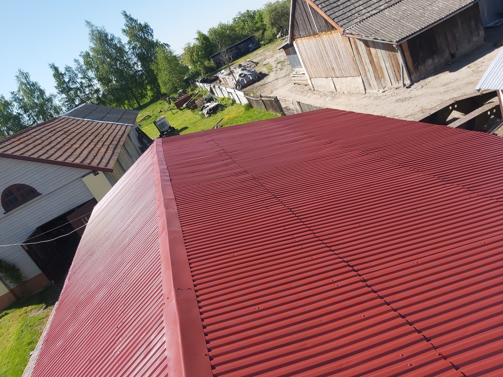 Czerwony, blaszany dach trapezowy, widok z góry na budynki gospodarcze i zieleń w tle.