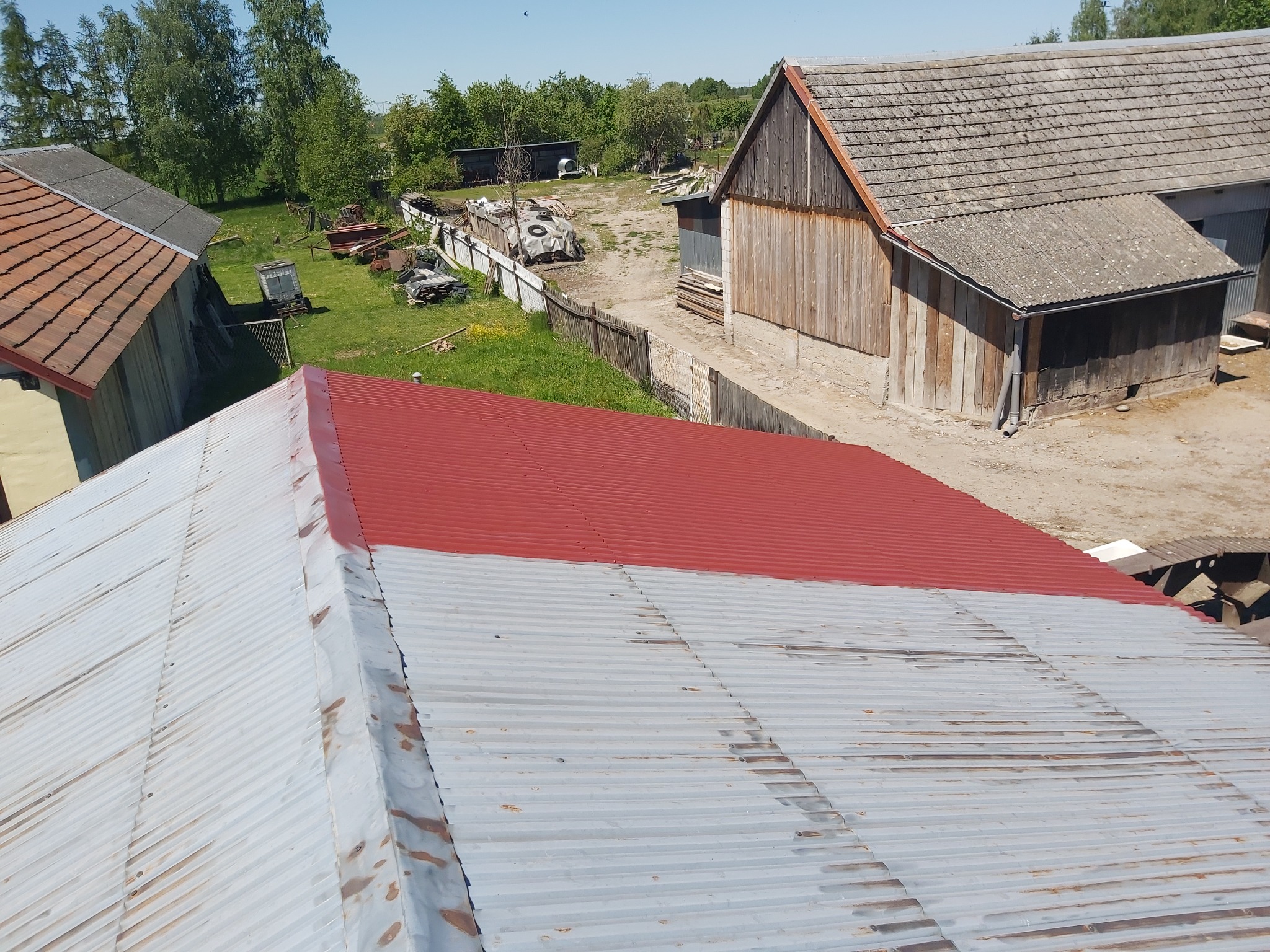 Częściowo pomalowany, blaszany dach budynku gospodarczego z widocznymi śladami rdzy, otoczony zabudowaniami gospodarczymi i zielenią w słoneczny dzień.