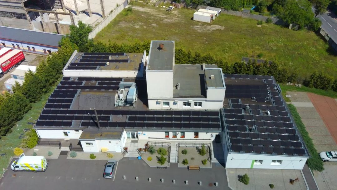 Widok z lotu ptaka na budynek z panelami słonecznymi na dachu, zaparkowanym samochodem osobowym i vanem firmy oferującej usługi związane z odnawialnymi źródłami energii, z zielenią w tle.