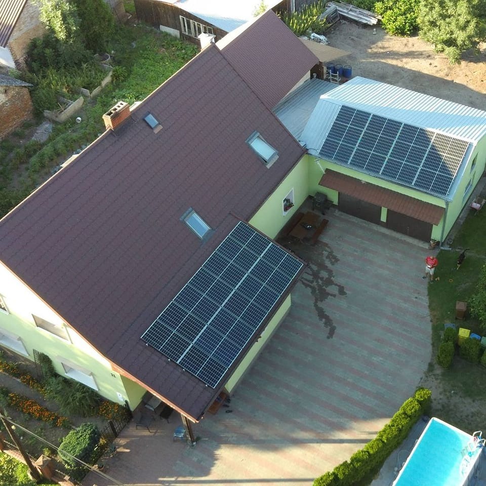 Widok z góry na dom z brązowym dachem i panelami słonecznymi na dachu oraz na budynku gospodarczym, obok basen i osoba z psem.