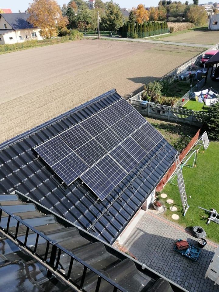 Panele słoneczne na dachu domu jednorodzinnego z czarną dachówką, widok z góry na instalację i otaczający krajobraz z polami i zielenią, drabina oparta o dach.