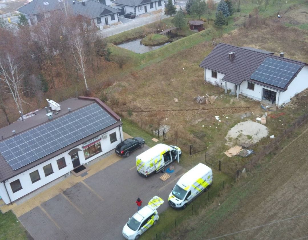 Widok z drona na budynki z panelami słonecznymi na dachach, zaparkowane samochody firmowe i wykopane rowy w ziemi.