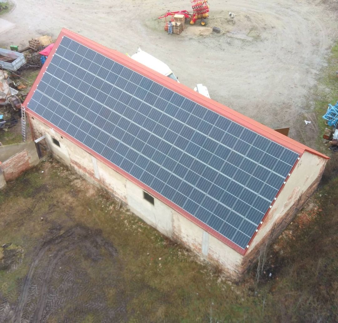 Widok z góry na duży budynek gospodarczy z panelami słonecznymi na dachu, obok zaparkowany biały samochód dostawczy, w tle sprzęt rolniczy.
