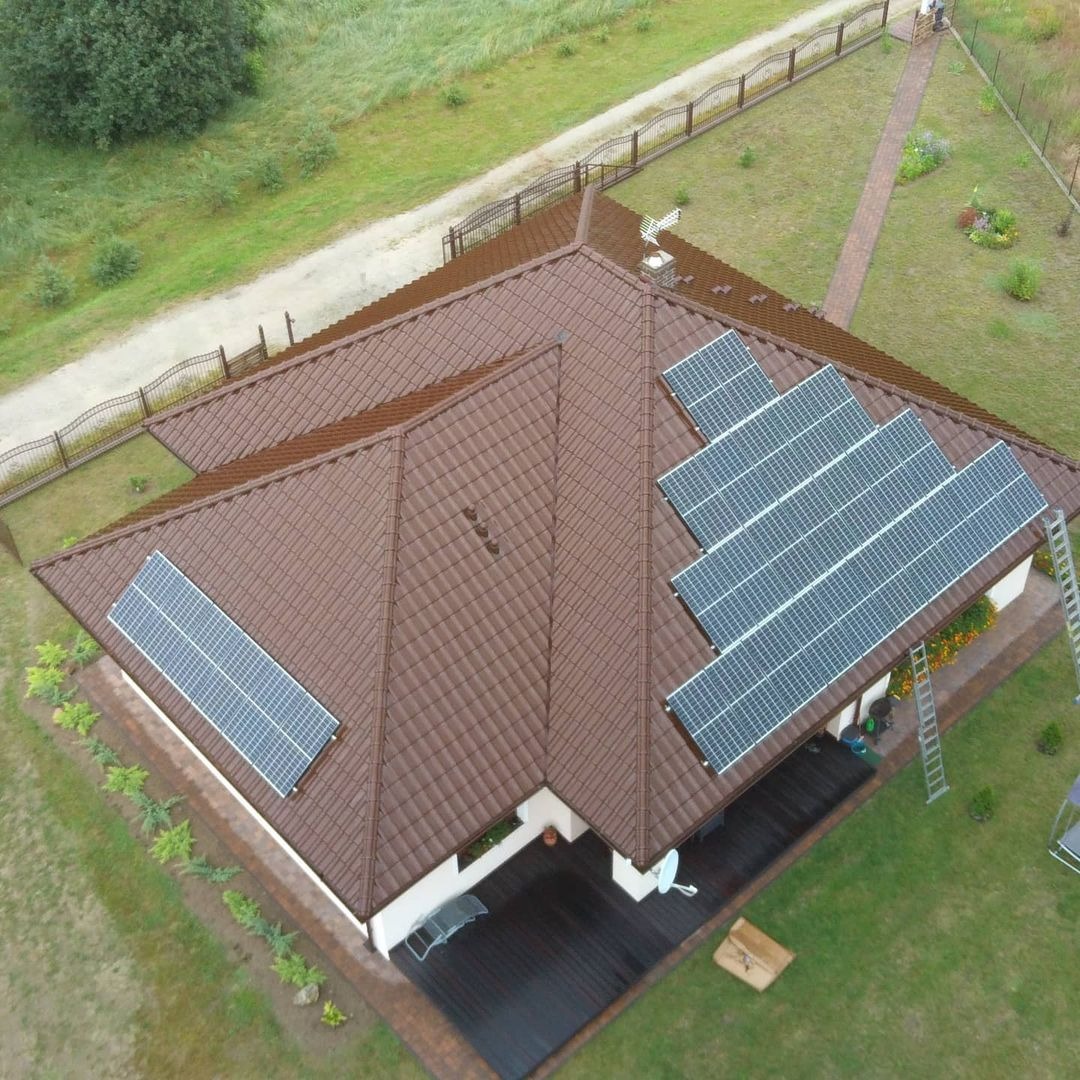 Widok z drona na brązowy dach domu z zainstalowanymi panelami słonecznymi; część paneli ułożona wzdłuż kalenicy, drabina oparta o dach, zadbany ogród.