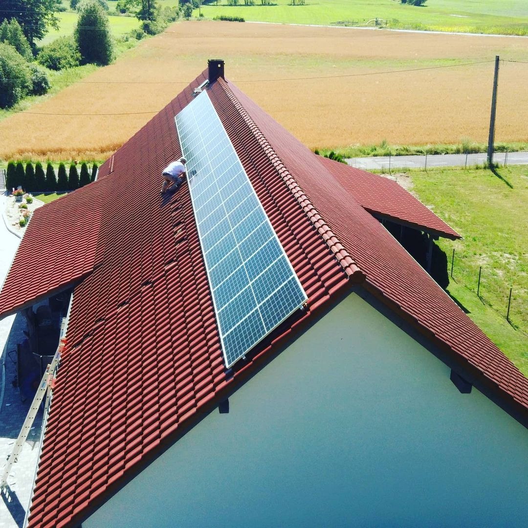 Montaż paneli słonecznych na dachu domu z czerwoną dachówką, widok z góry na instalatora pracującego na dachu, w tle pole uprawne.