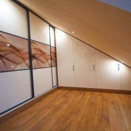 MEBLE GAL Tadeusz Gal - Szafa wnękowa z przesuwnymi drzwiami z abstrakcyjnym wzorem w odcieniach brązu, dopasowana do skosu poddasza, z drewnianą podłogą w tle.