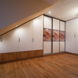 MEBLE GAL Tadeusz Gal - Szafa wnękowa z przesuwnymi drzwiami z grafiką w brązowych odcieniach, zabudowa pod skosem z białymi frontami i drewnianymi szufladami, oświetlenie punktowe, parkiet.