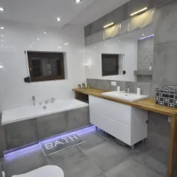 Nowoczesna łazienka w szaro-białej kolorystyce z drewnianym blatem, podświetlaną wanną i minimalistycznym grzejnikiem drabinkowym.