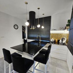 MEBLE GAL Tadeusz Gal - Nowoczesna kuchnia w bieli i czerni z wyspą, czarnym blatem z połyskiem i złotymi akcentami w postaci baterii i lamp wiszących.