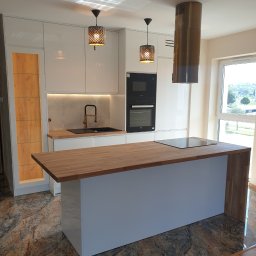 MEBLE GAL Tadeusz Gal - Nowoczesna kuchnia z wyspą, białe lakierowane fronty, drewniany blat, okrągły okap w kolorze mosiądzu, dekoracyjne lampy wiszące, marmurowe płytki podłogowe.